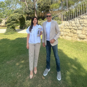 A man and woman stand together in front of a textured stone wall, both smiling and dressed casually.